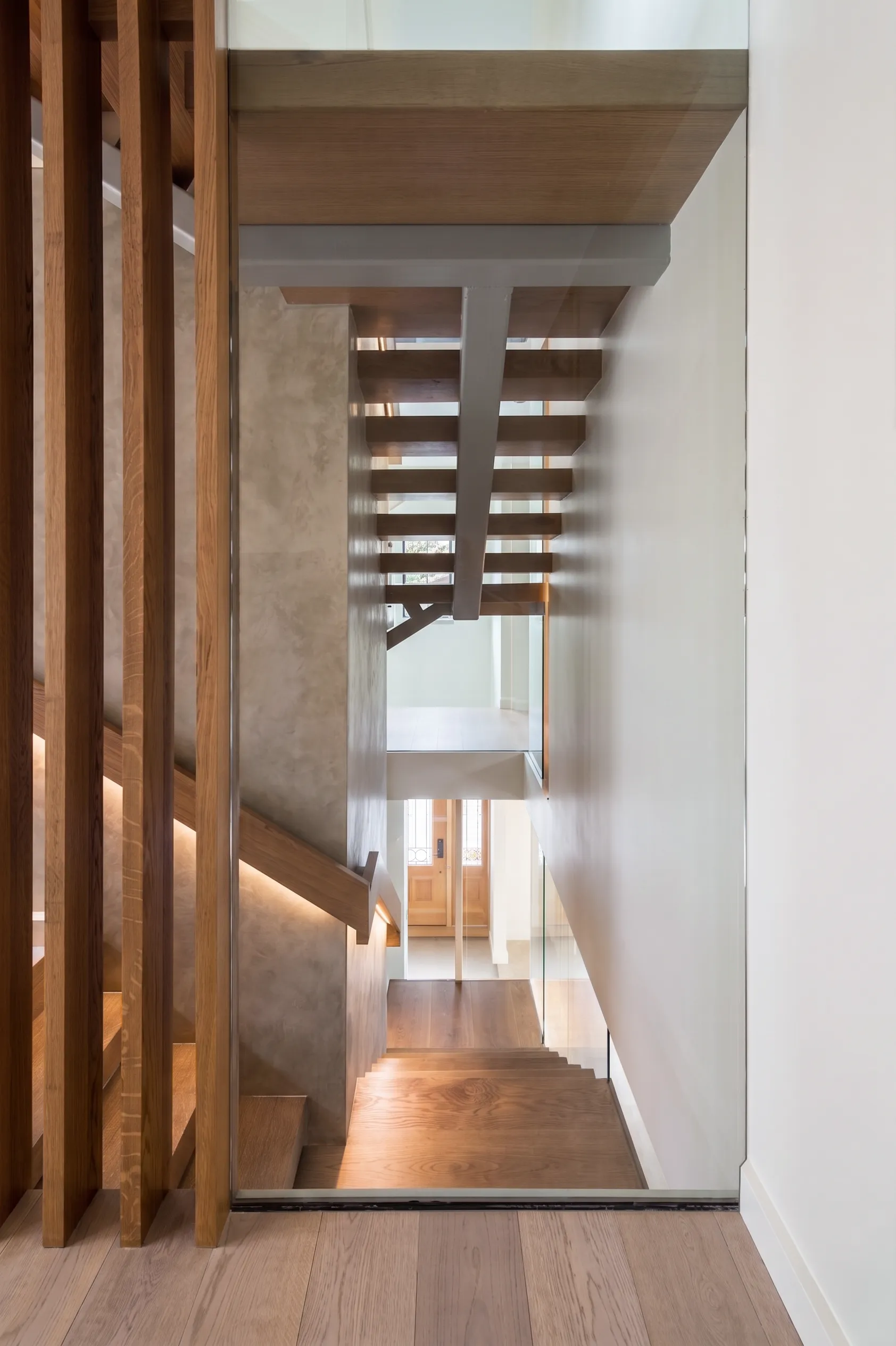 Floating wood stair wrapped in glass descending past a plaster wall toward the entry of a Long Island multigenerational home