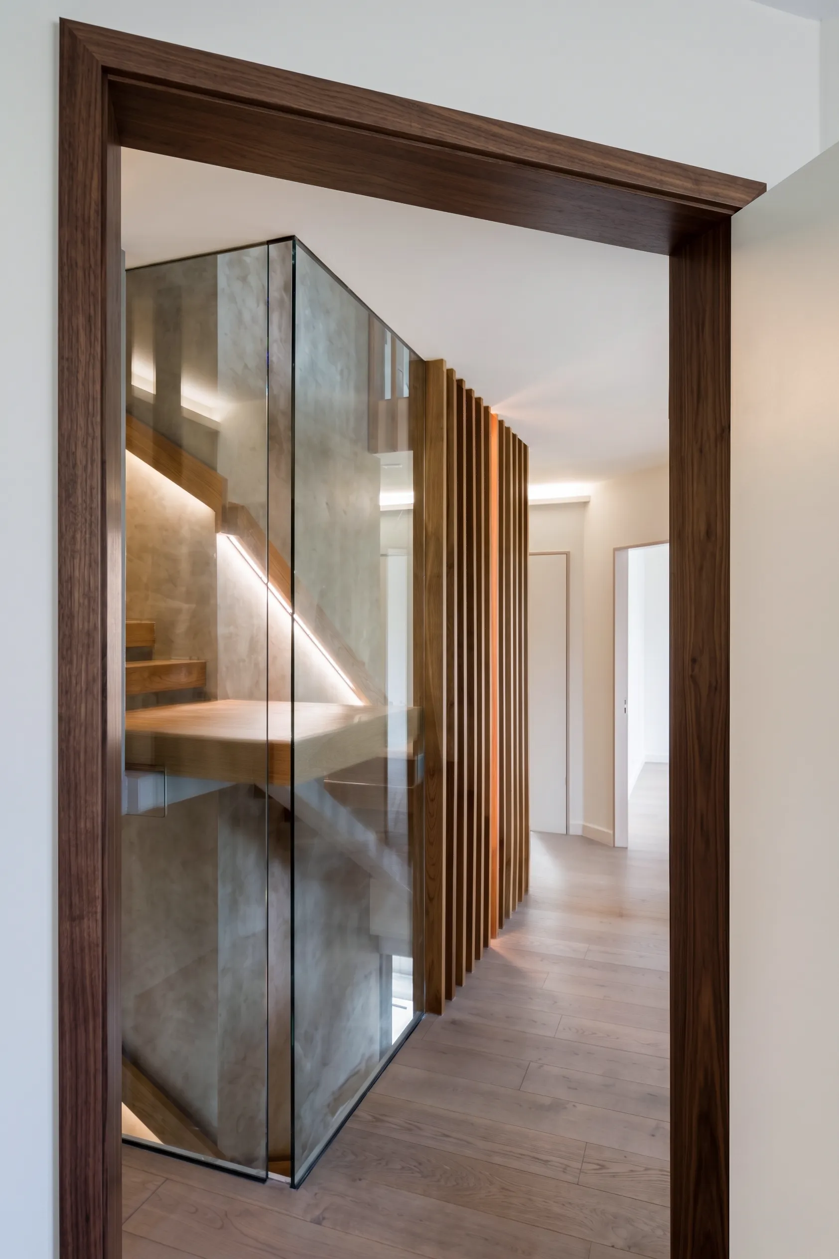 Walnut-framed doorway opening to a glass-wrapped sculptural stair and wood-slat screen in a Long Island home renovation