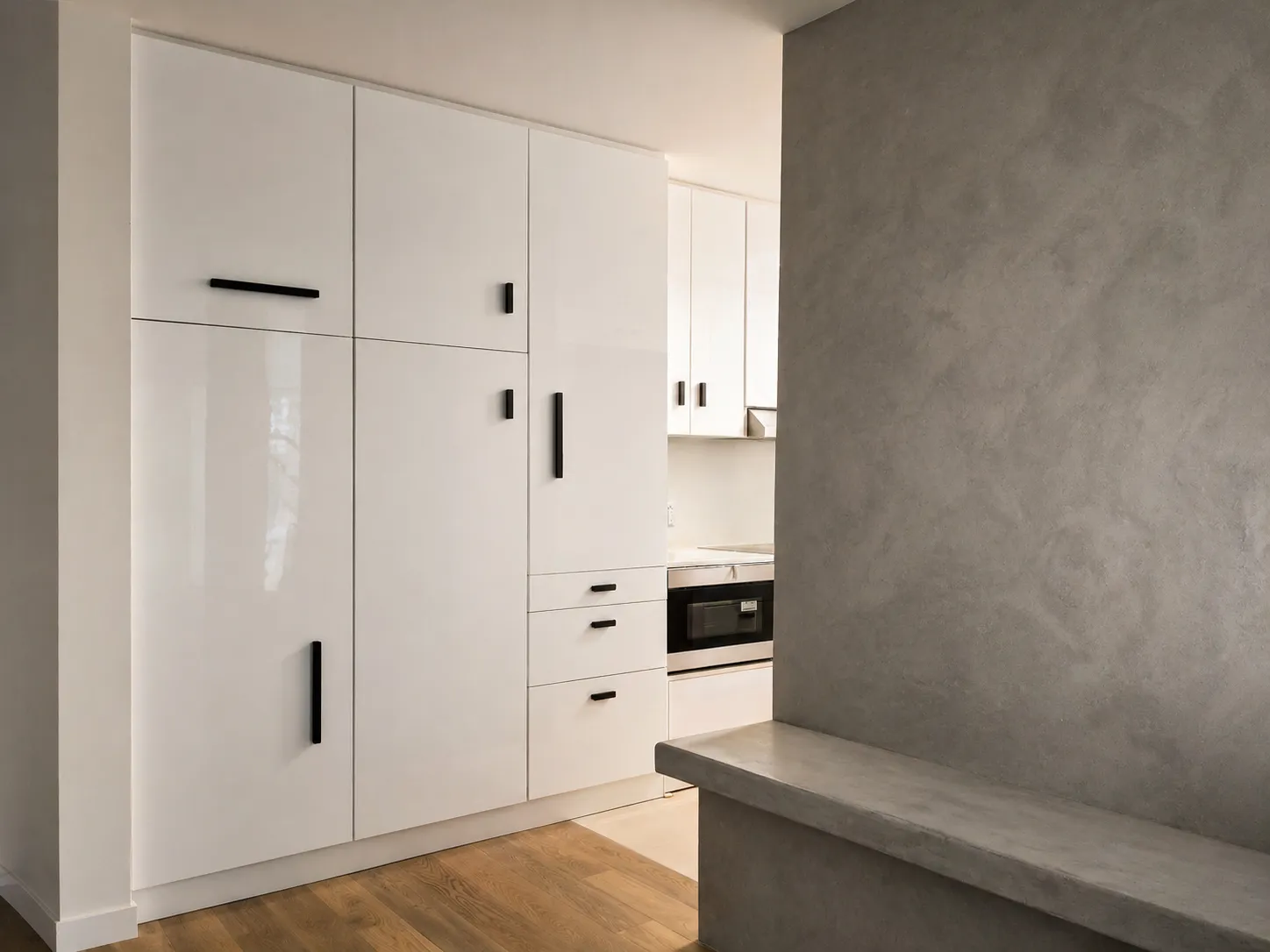 Floor-to-ceiling white gloss cabinetry with black bar pulls beside a plaster wall and concrete bench in a Manhattan apartment kitchen