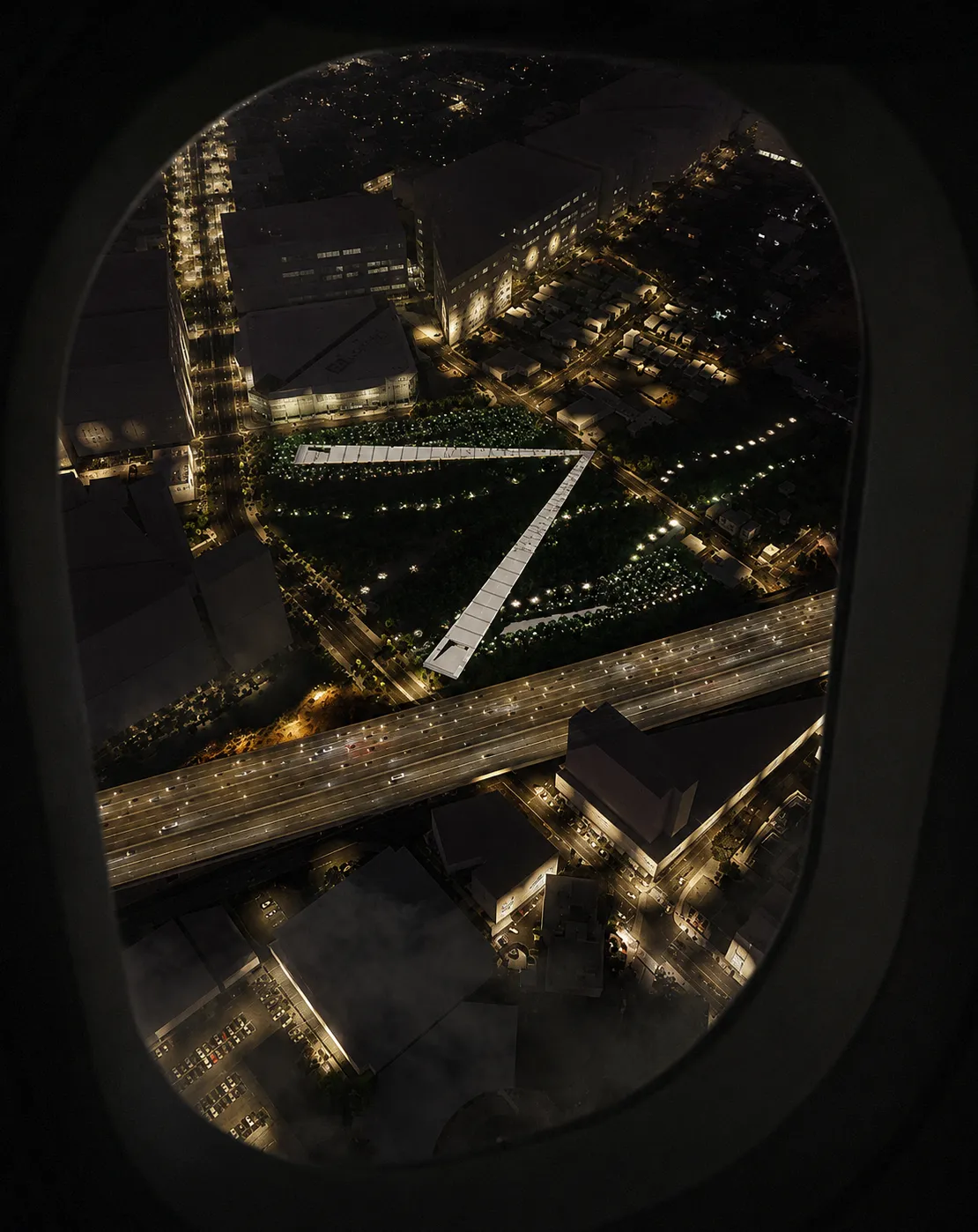 Night aerial through an airplane window of the lit walkway forming an arrow across a dark San Jose park beside a lit highway