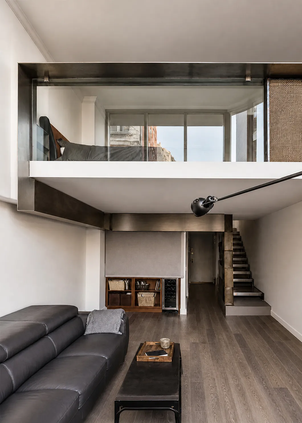 Double-height living room of an Upper West Side duplex with a gray leather sofa below and a steel-and-glass mezzanine bedroom above