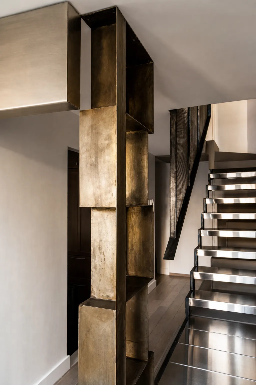 Blackened steel open-tread stair with a woven metal mesh guard in a Manhattan duplex, set beside a brass box-shelving column