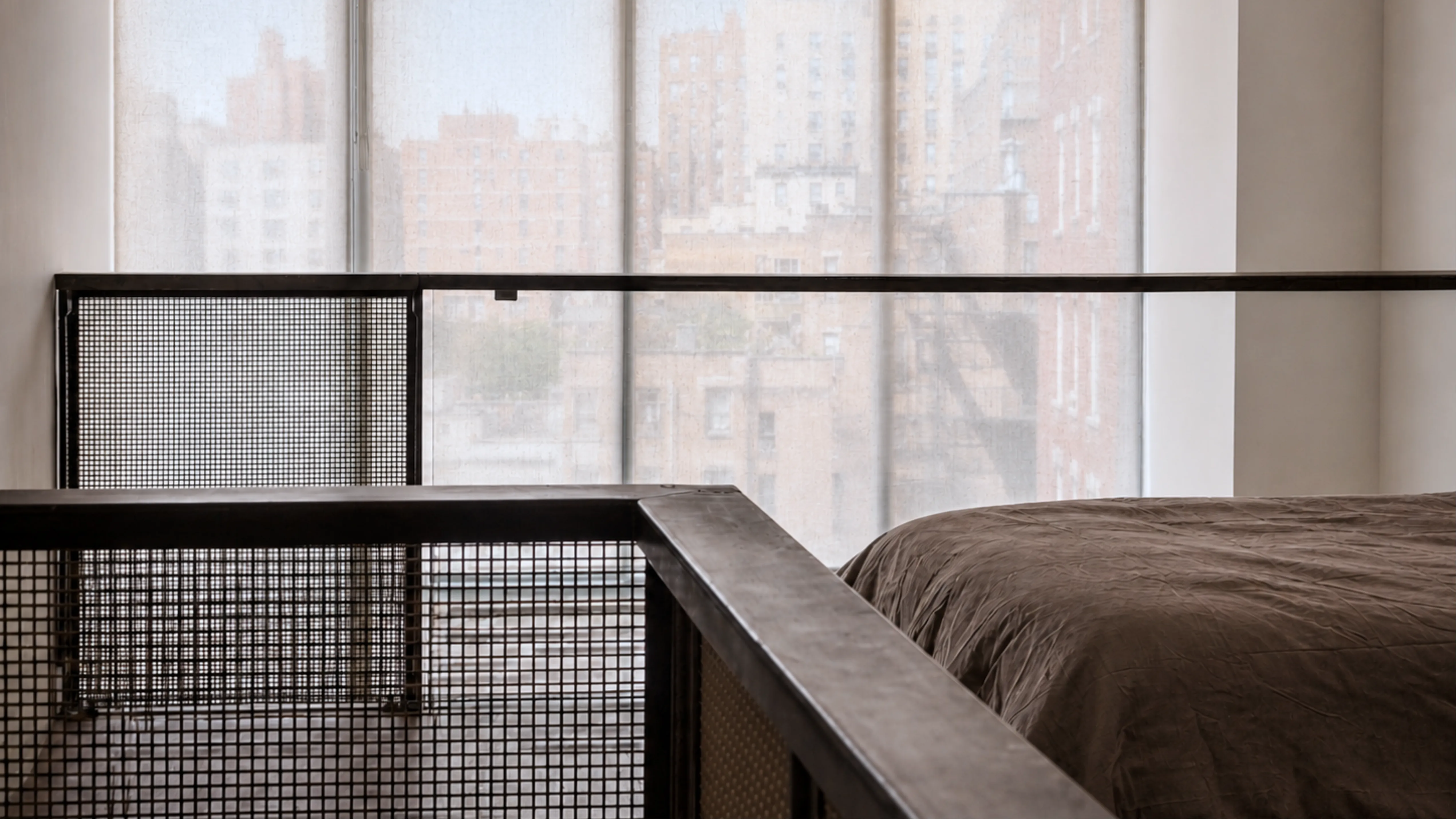 Mezzanine bed behind a blackened steel railing with metal mesh infill, framed by soft white window shades in a Manhattan duplex