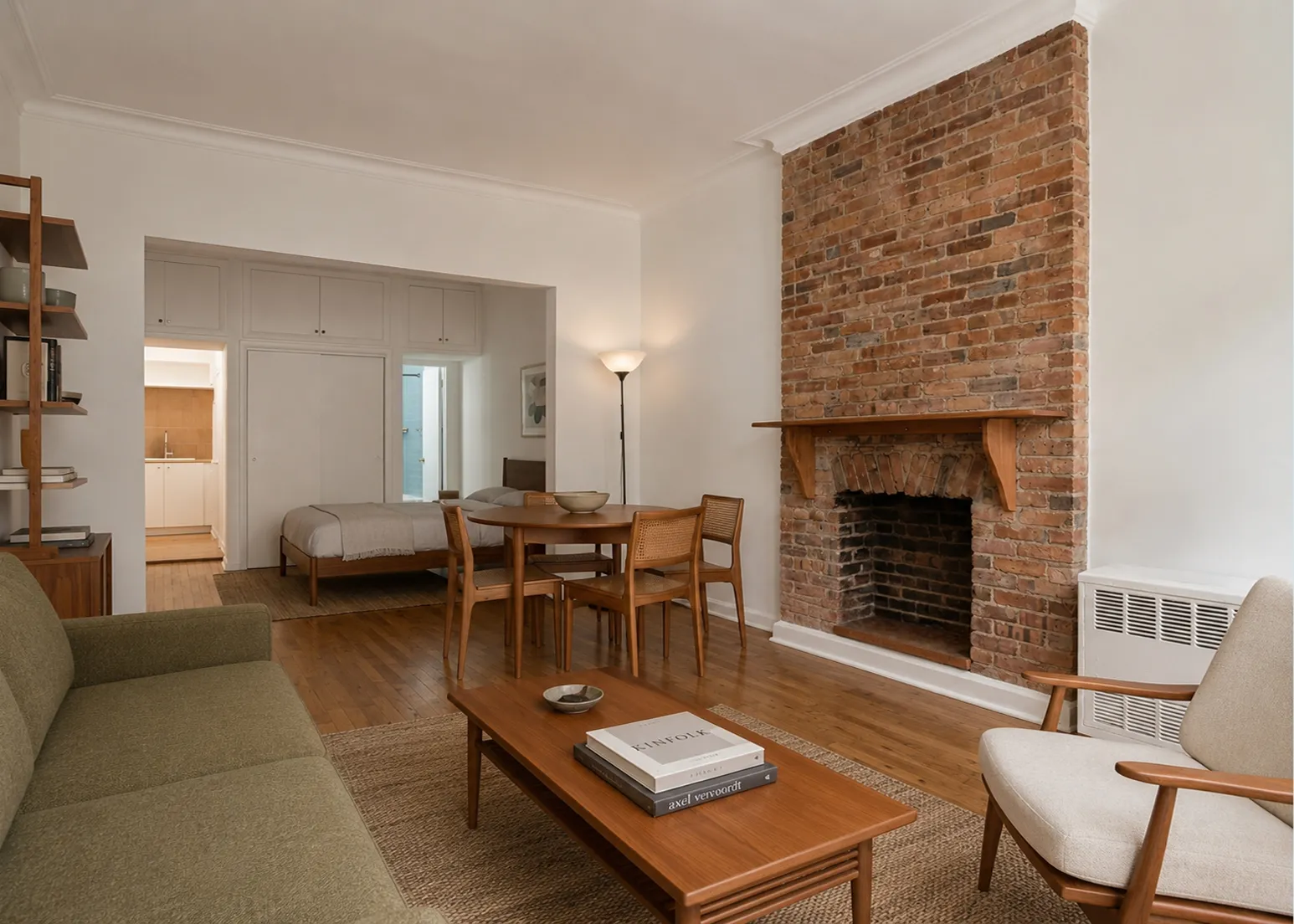 Living area of a Prospect Heights studio with a green sofa and walnut coffee table beside an exposed brick fireplace, with the bed beyond