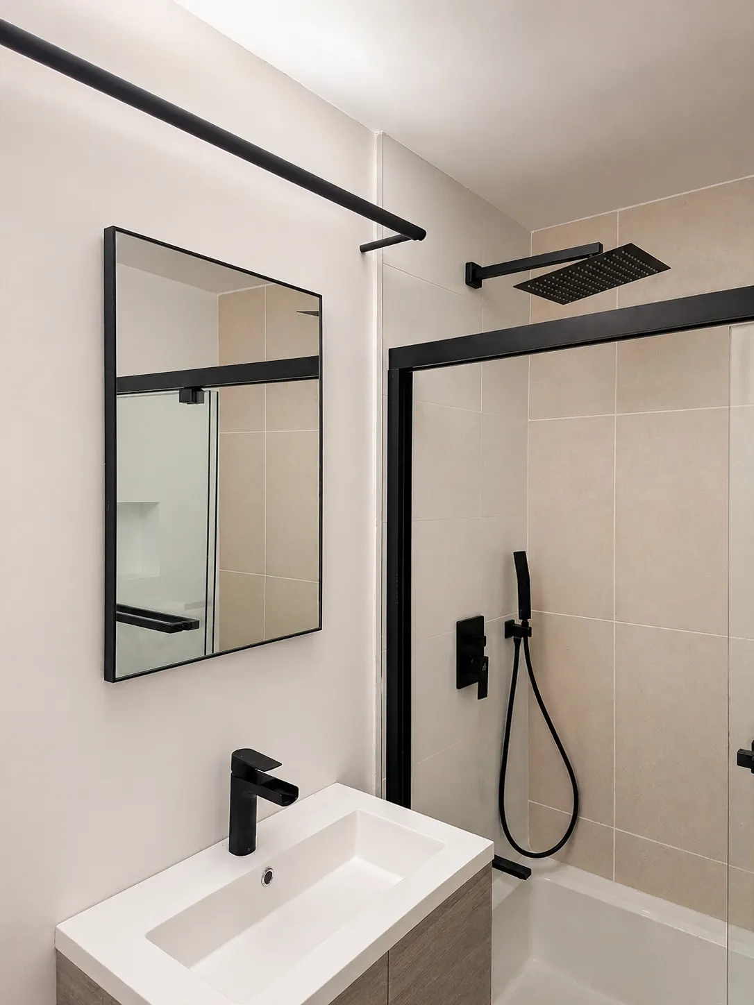 Compact Manhattan apartment bathroom with matte black fixtures, a black-framed mirror, and a beige-tiled shower behind a black-framed glass door