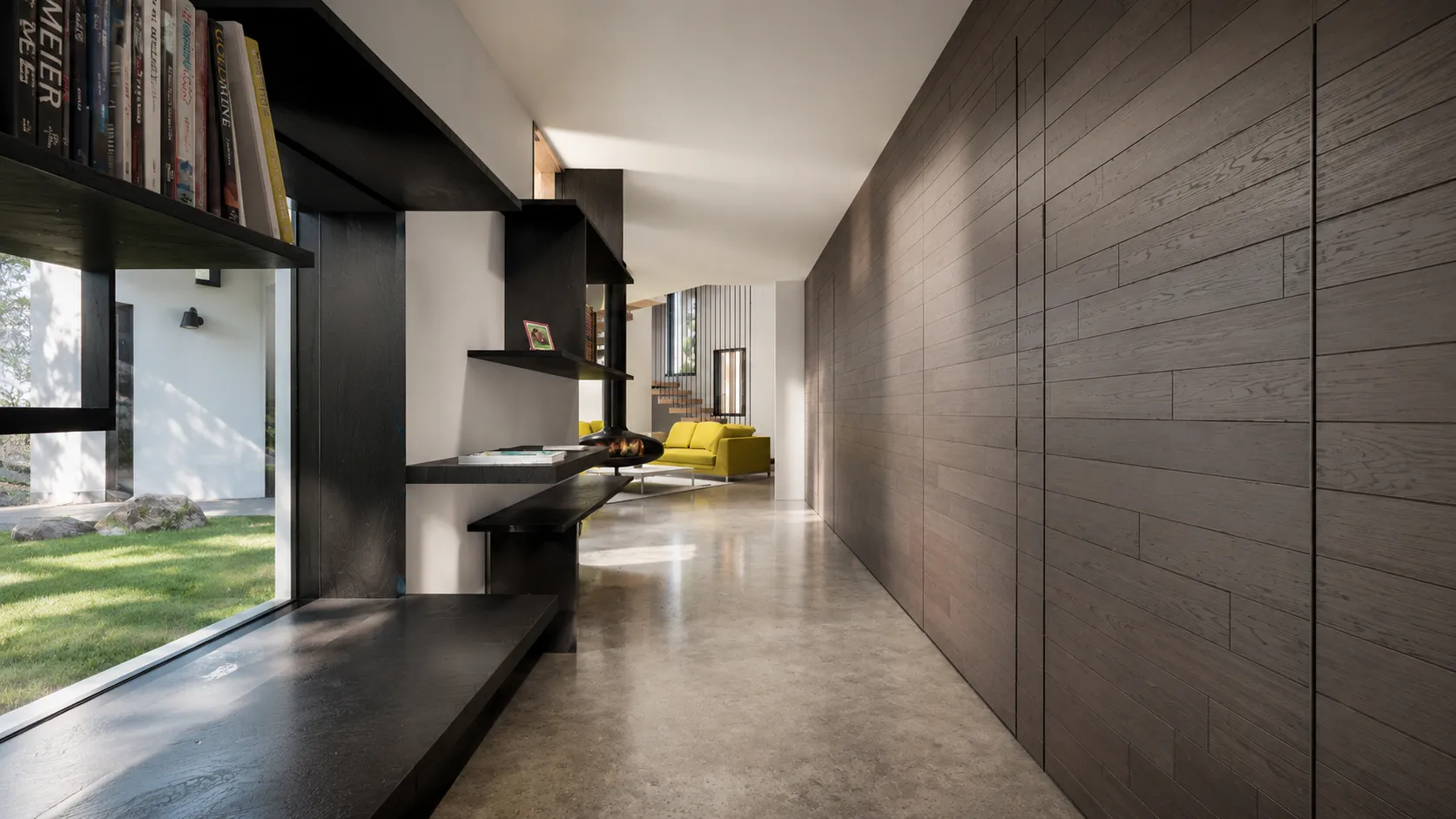 Gallery hall in a Staatsburg forest house with blackened-wood built-in shelves along a window wall and a polished concrete floor