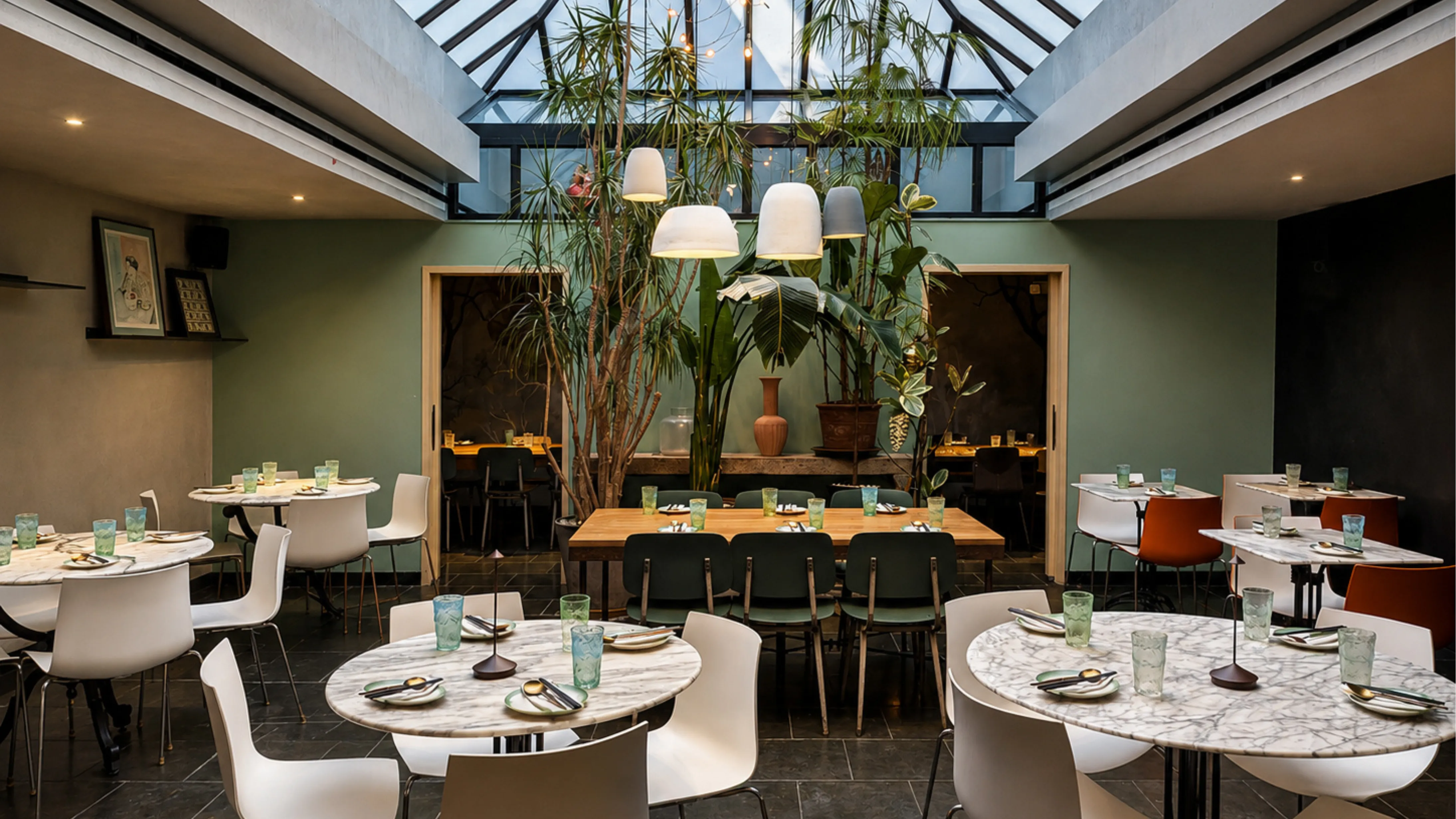 Noodle restaurant dining room with a glass skylight, green walls, tall indoor plants, and marble café tables in Chelsea, New York