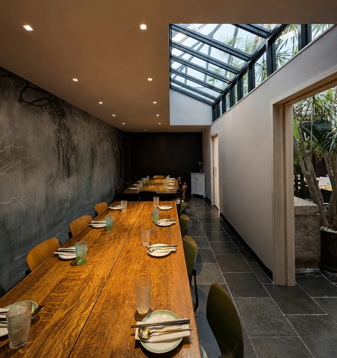 Private dining room with a long wood communal table, charcoal tree mural, and skylight in a noodle restaurant in Chelsea, New York