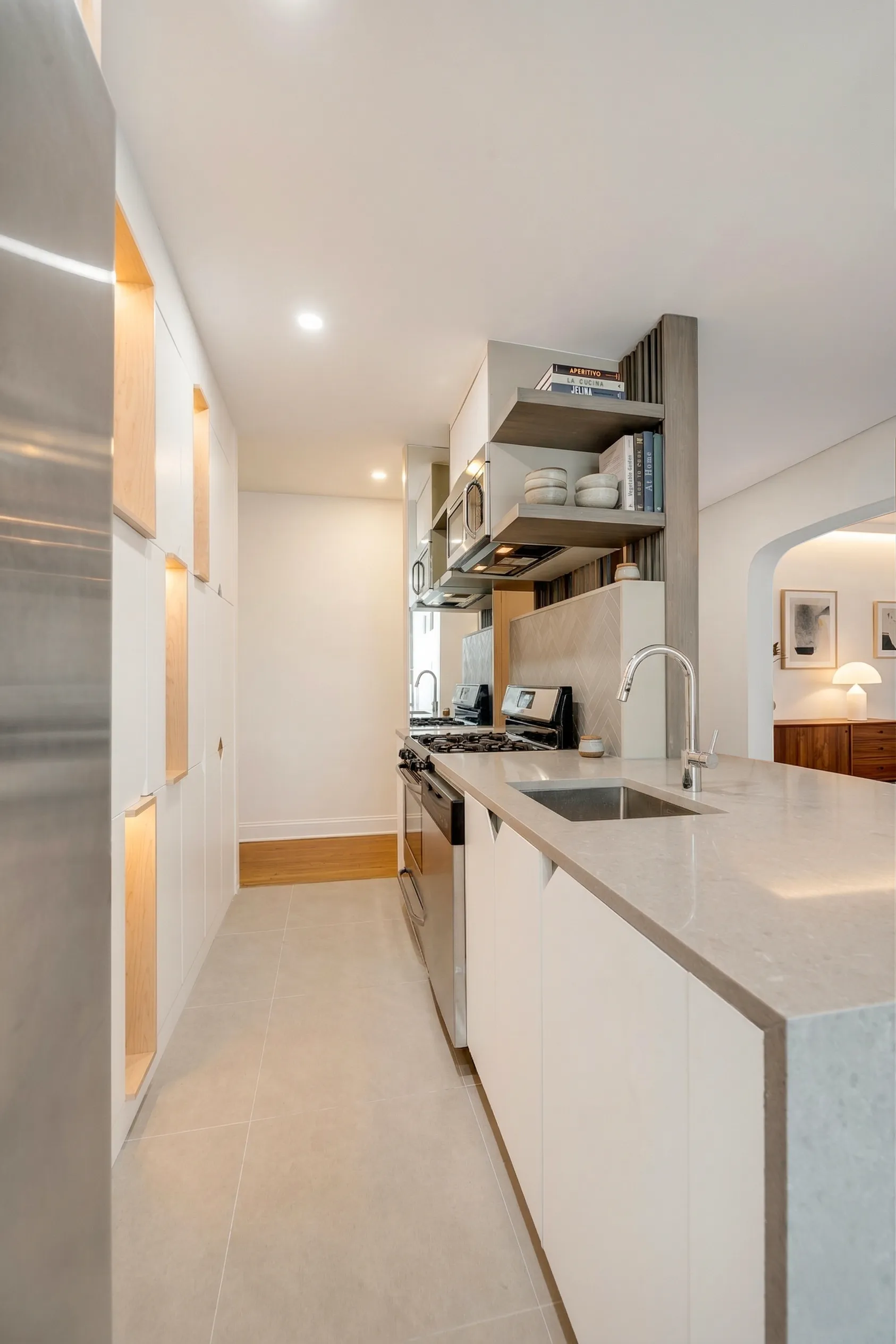 Galley kitchen in a Brooklyn apartment with white cabinetry, wood-lined niches, a stone peninsula, and an arched opening to the dining room