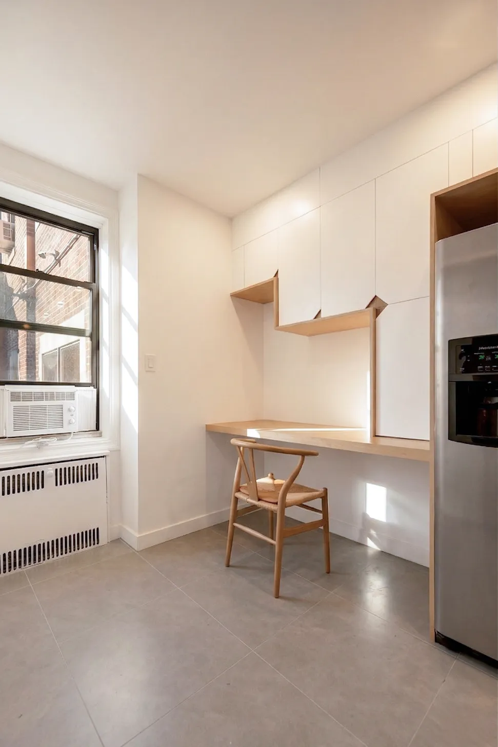 Sunlit breakfast nook in a Brooklyn kitchen with a built-in wood desk, a wishbone chair, and white cabinets beside a stainless fridge