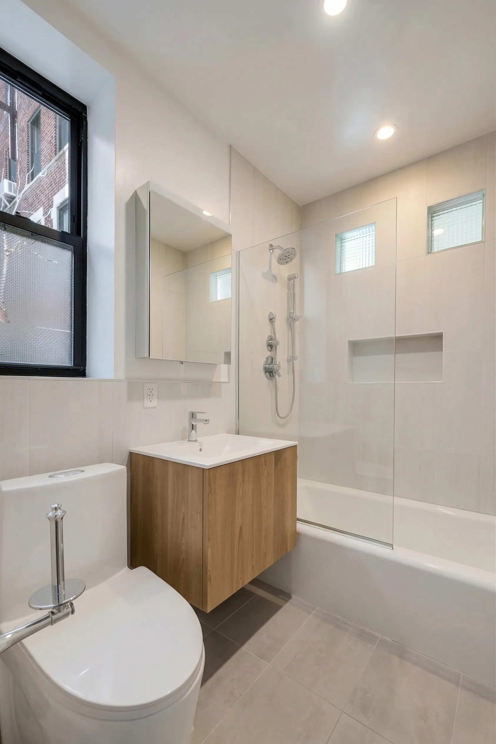 Brooklyn bathroom with cream tile, a wood floating vanity, a medicine-cabinet mirror, and a glass-paneled tub shower