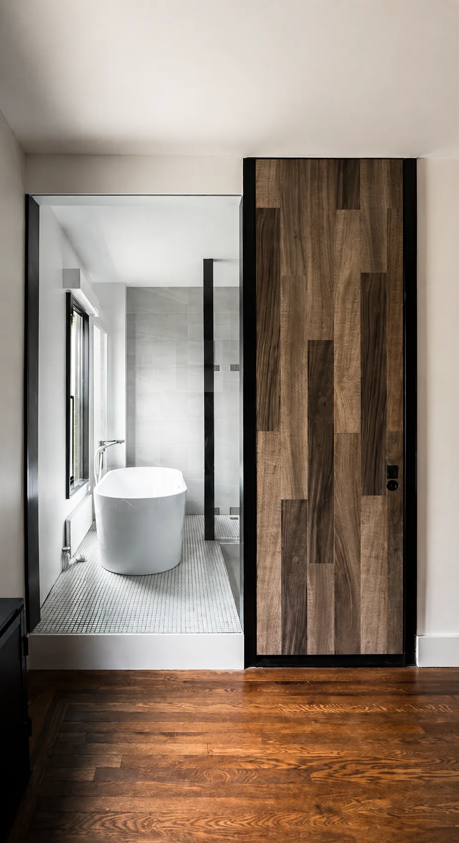 Wood-plank pivot door opening to a Manhattan master bath with a freestanding white tub beside a window and a mosaic tile floor