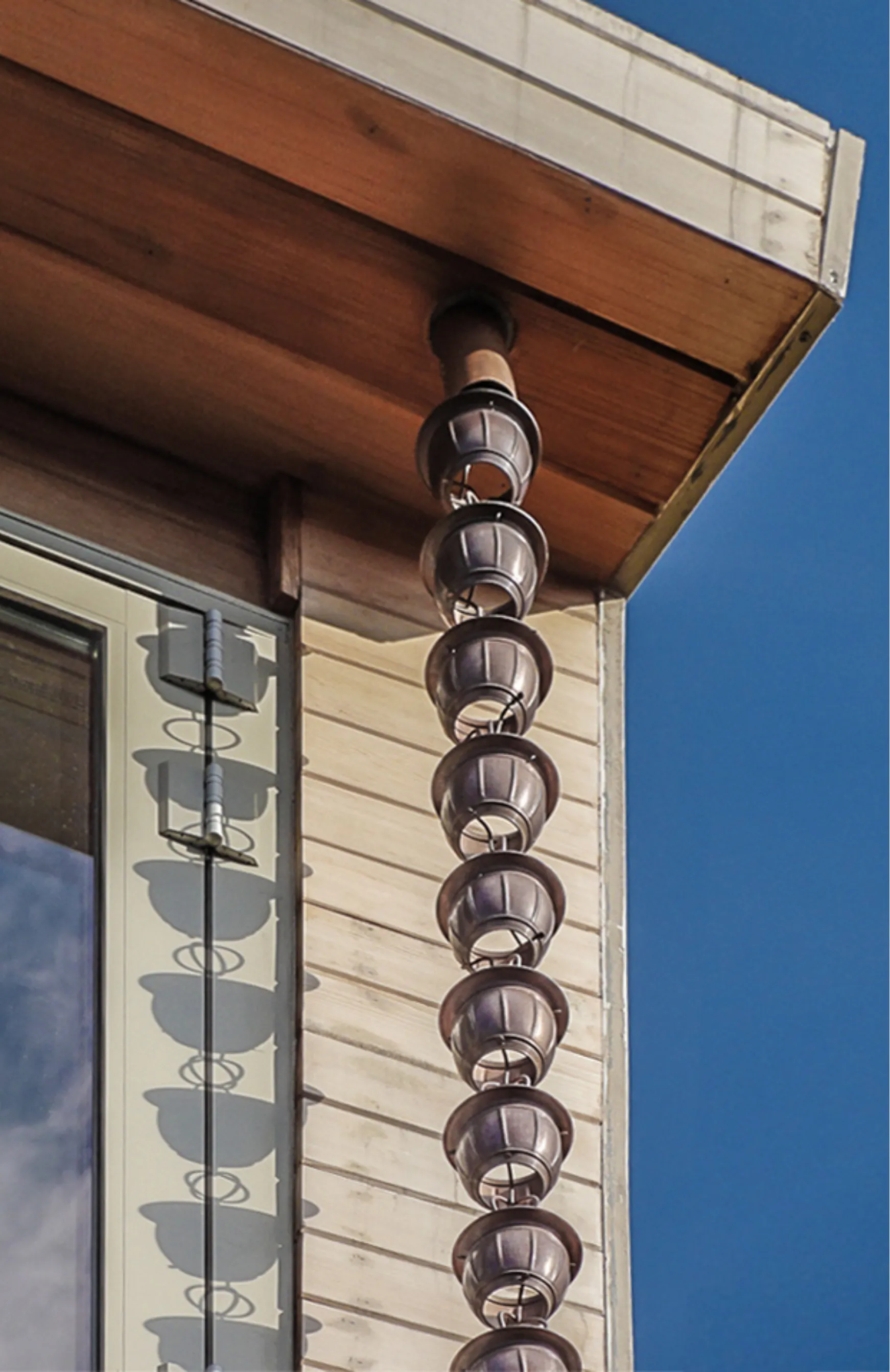 Copper-cup rain chain hanging from a cedar soffit on a Park Slope balcony sunroom, casting bowl-shaped shadows across white clapboard siding